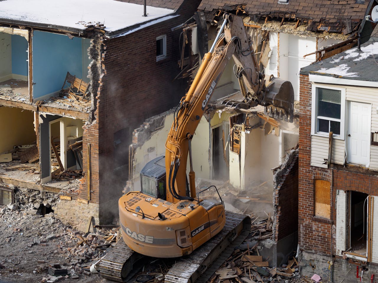 services-02 An excavator demolishes an old brick building at a construction site, creating debris and dust.